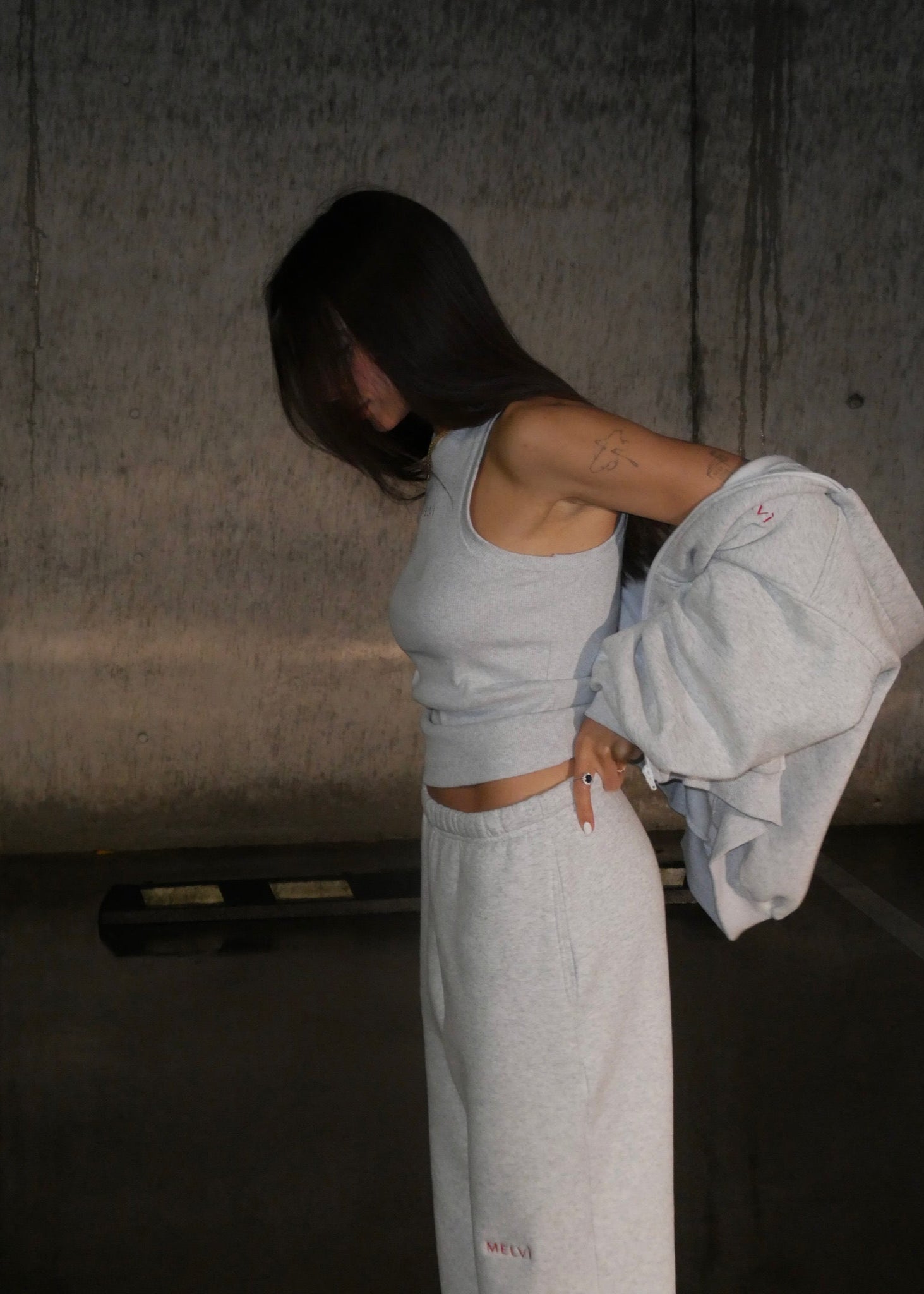Person wearing a light gray tank top and pants, holding a jacket in a dimly lit room.