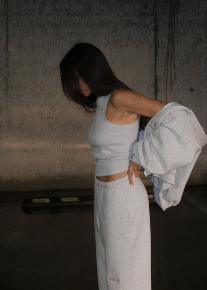 Person wearing a light gray tank top and pants, holding a jacket in a dimly lit room.